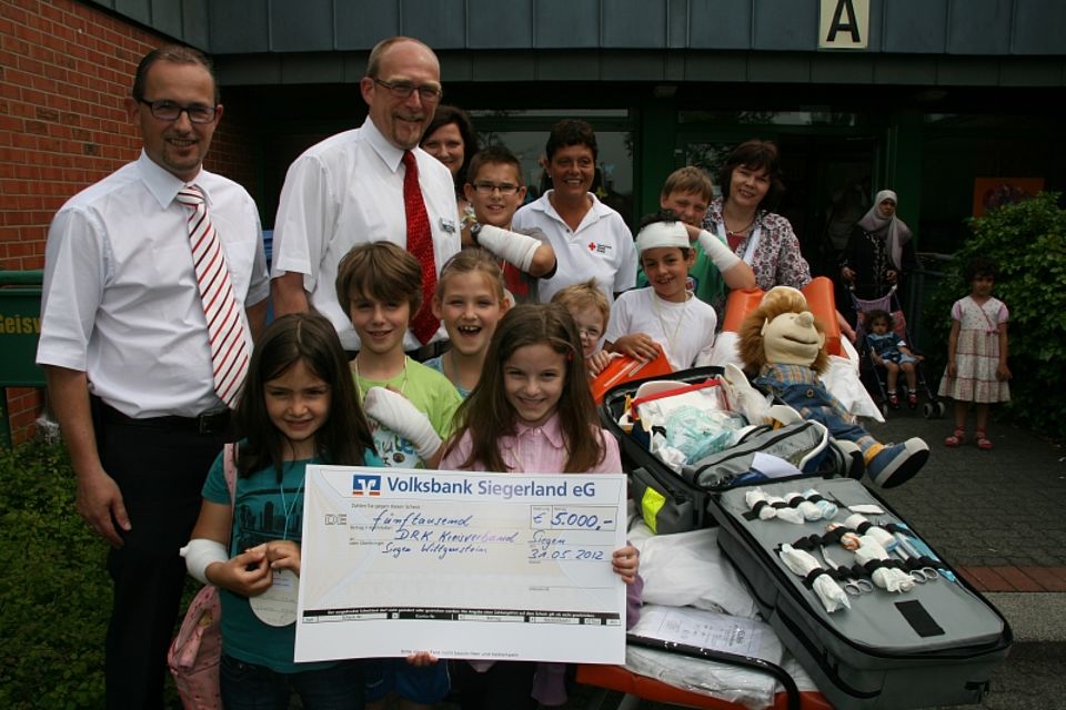 v.l. Jörg Hoffmann (Volksbank Siegerland eG) übergab Ralf Henze (DRK-Kreisgeschäftsführer) im Beisein von,Petra Trogisch (DRK-Ausbilderin), Silvia Wetter-Wicht (Schulleiterin Geisweider Schule) und der kleinen Geisweider Grundschüler, den Scheck über 5000 Euro.